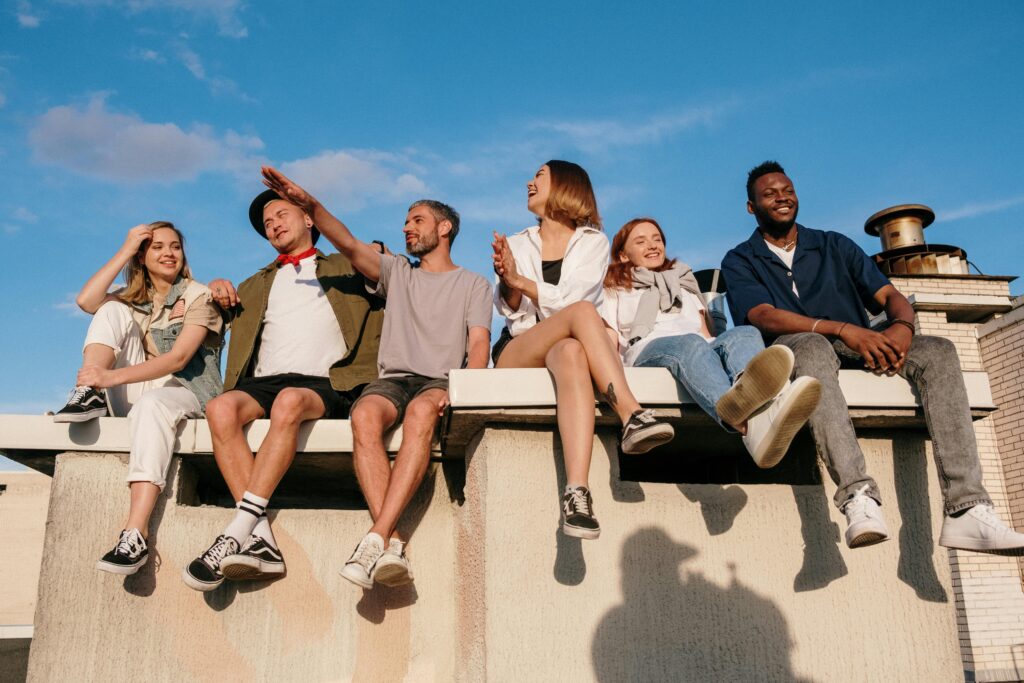 A diverse group of friends laughing and enjoying a sunny day on a rooftop with a blue sky background.