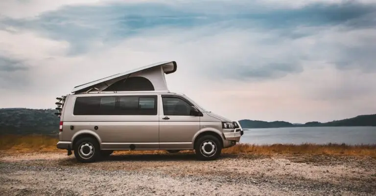 A camper van with rooftop tent parked by a tranquil lake under cloudy skies, ideal for adventure travel.